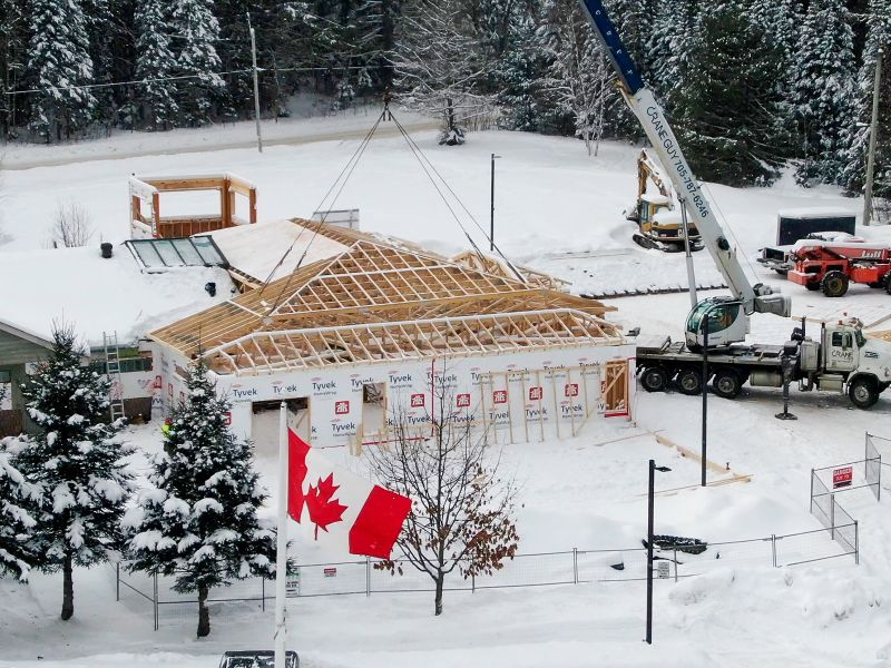 A crane lifts a large wooden roof section into place on the snowy construction site.